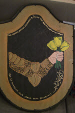 Device: Sable, a sinister arm fesswise reversed embowed proper armored Or grasping three tulips Or slipped vert all within a bordure Or.

About the additional text on my shield: I have always liked the song 'Tub-Thumping' by Chumbawamba. While it is about politicians who are 'stumping' (The term used here in America) for election I took it as a personal theme song when my health tanked a few years ago. So with a bit of help from Google and Babel translations on-line I give you:

Ego Adepto infligo d